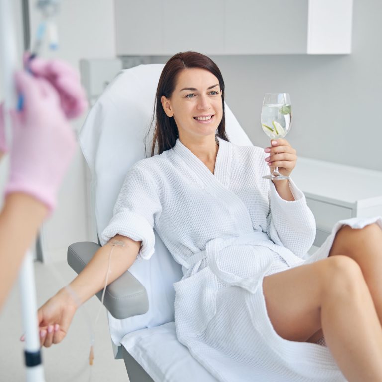 Smiling good-looking dark-haired spa client relaxing with a glass of beverage in a cosmetic chair